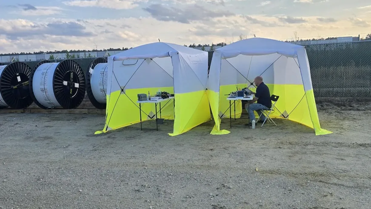 Two work tents on gravel next to cable reels, a technician seated at a table inside.